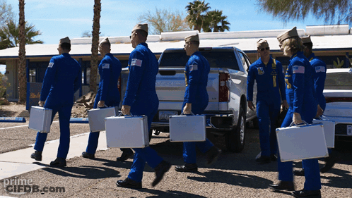A Group Of Men In Blue Jumpsuits Are Carrying Briefcases In Front Of A Silver Truck That Says Gmc On The Back GIF