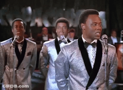 A Group Of Men In Tuxedos Are Singing Into Microphones . GIF