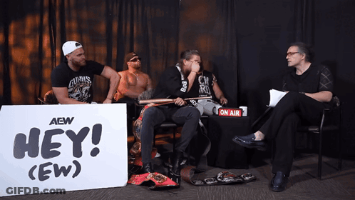 A Group Of Men Sitting In Front Of A Sign That Says Hey GIF