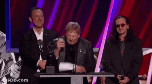 A Group Of Men Standing Around A Podium With One Wearing Glasses GIF