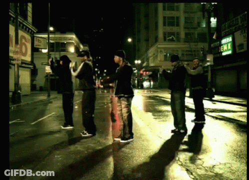 A Group Of Men Standing On A Wet Street In Front Of A Sign That Says Emm GIF