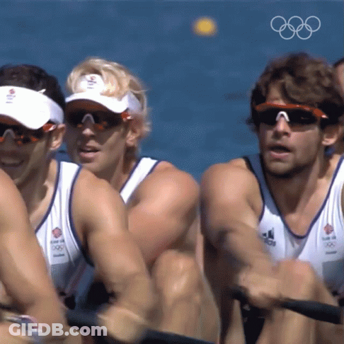 A Group Of Men Wearing Sunglasses And Visors Are Rowing In The Water With The Olympic Rings In The Background GIF