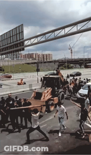 A Group Of People Are Carrying A Large Piece Of Wood In Front Of A Bulldozer On A Highway GIF