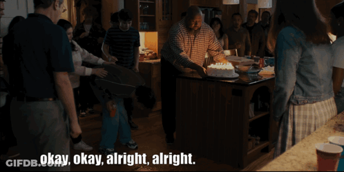 A Group Of People Are Gathered Around A Man Holding A Cake With The Words Okay Okay Alright Alright Below Him GIF