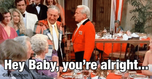 A Group Of People Are Gathered Around A Table With A Man In A Red Jacket Saying Hey Baby You 'Re Alright GIF