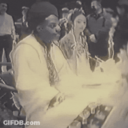 A Group Of People Are Gathered In A Dark Room And One Of Them Is Wearing A Black Hat GIF