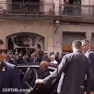 A Group Of People Are Gathered Outside A Building With A Blue Sign On The Wall That Says ' A ' On It GIF