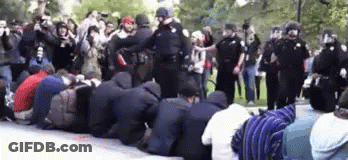 A Group Of People Are Kneeling Down In Front Of A Police Line GIF