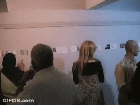 A Group Of People Are Looking At Pictures On A Wall And One Woman Is Pointing At One Of Them GIF