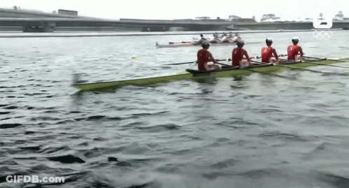 A Group Of People Are Rowing A Boat On A Lake With The Olympic Logo In The Background GIF