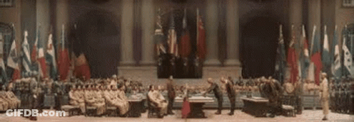 A Group Of People Are Sitting Around A Table In A Large Room With Flags Behind Them . GIF