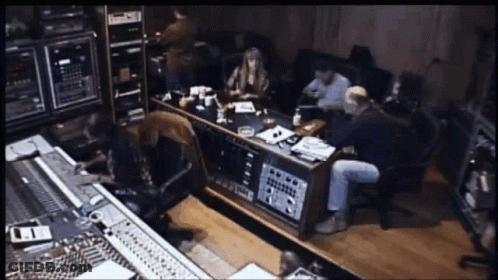 A Group Of People Are Sitting Around A Table In A Recording Studio With A Sign On The Wall That Says ' A ' GIF