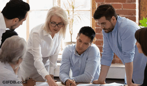 A Group Of People Are Sitting Around A Table Looking At A Piece Of Paper . GIF