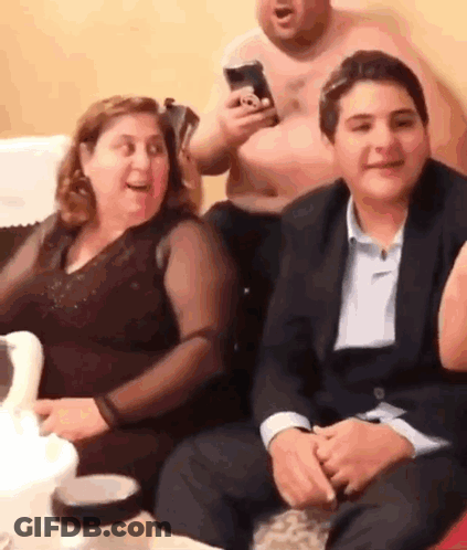 A Group Of People Are Sitting On A Couch And Laughing While A Man Holds A Cell Phone In His Hand . GIF