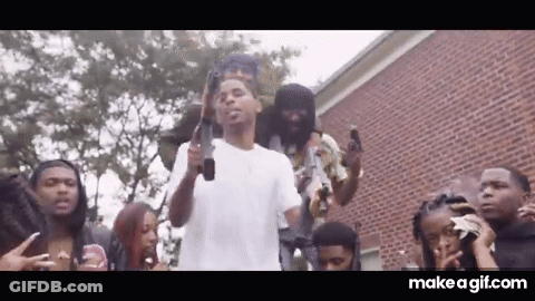 A Group Of People Are Standing Around A Man Holding A Gun In Front Of A Brick Building . GIF