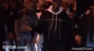 A Group Of People Are Standing Around A Man In A Black Jacket . GIF