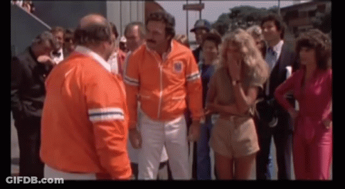 A Group Of People Are Standing Around A Man Wearing An Orange Jacket That Says ' New York ' On It GIF