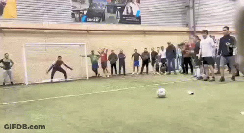 A Group Of People Are Standing Around A Soccer Field Watching A Goalie Kick A Soccer Ball . GIF