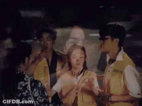 A Group Of People Are Standing In A Dark Room With A Man 'S Face Projected On A Wall Behind Them . GIF