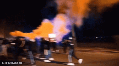A Group Of People Are Walking Down A Street At Night With Smoke Coming Out Of A Fire . GIF