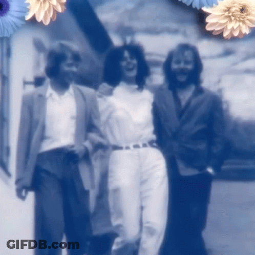 A Group Of People Are Walking Down A Street With Blue Flowers In The Background GIF