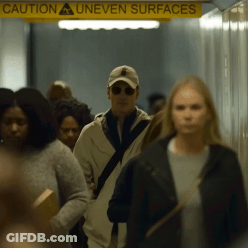 A Group Of People Are Walking In A Hallway Under A Yellow Sign That Says Caution Uneven Surfaces GIF