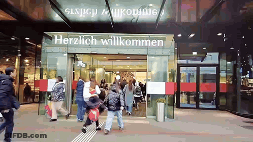 A Group Of People Are Walking In Front Of A Store That Says Herzlich Willkommen GIF
