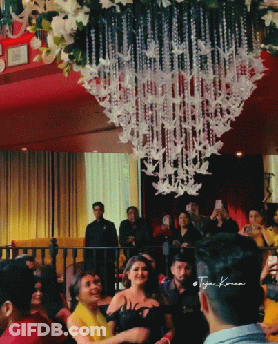 A Group Of People Gathered In A Room With A Chandelier Hanging From The Ceiling And A Man Taking A Picture GIF