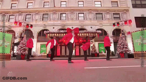 A Group Of People In Red Jackets Are Dancing In Front Of A Building With Christmas Trees . GIF