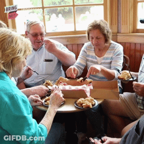 A Group Of People Sit Around A Table Eating Food With A Sign That Says Bring Me GIF