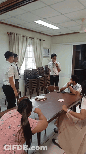 A Group Of People Sitting Around A Table With A Man Standing Behind Them Talking GIF
