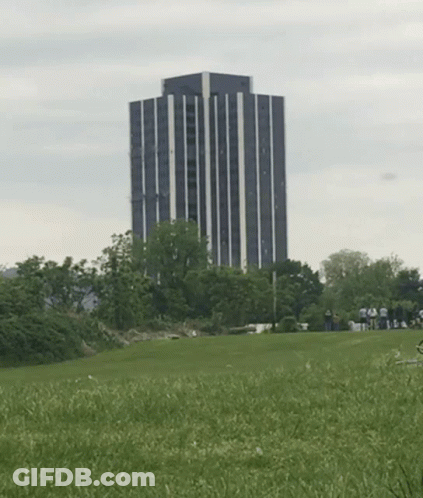 A Group Of People Standing In A Grassy Field With Smoke Coming Out Of A Building In The Background GIF