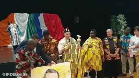 A Group Of People Standing In Front Of A Stage With A Man In A Yellow Robe Holding A Painting GIF