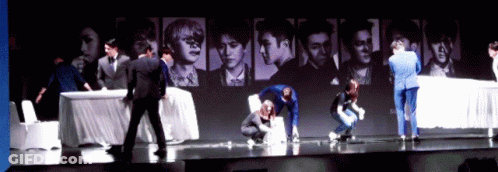 A Group Of People Standing On A Stage In Front Of A Wall With Pictures Of People GIF