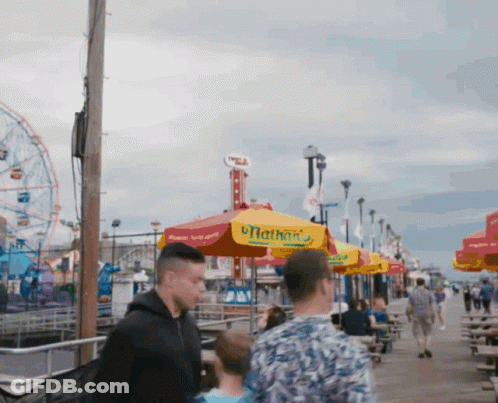 A Group Of People Walking Down A Boardwalk With Umbrellas That Say Hot Dogs GIF
