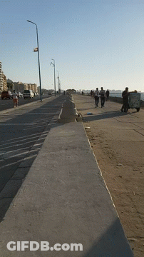 A Group Of People Walking Down A Sidewalk With A Trash Can On The Side GIF