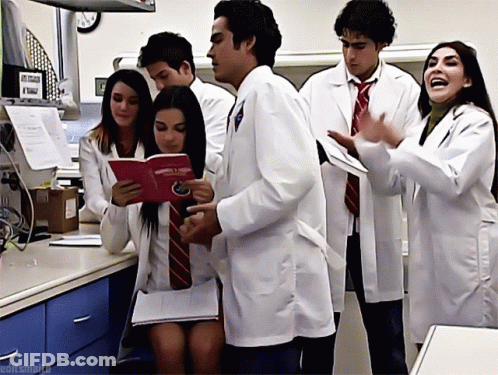 A Group Of People Wearing Lab Coats And Ties Are Standing Around A Table And One Of Them Is Holding A Book GIF