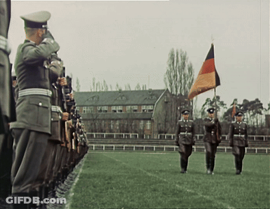 A Group Of Soldiers Marching In A Field With A Flag In The Background GIF