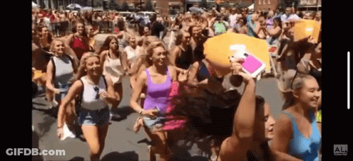 A Group Of Women Are Running Down A Street With A Man Holding A Pillow That Says Al On It GIF