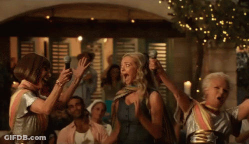 A Group Of Women Singing Into Microphones In A Crowd GIF