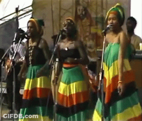 A Group Of Women Singing Into Microphones While Wearing Colorful Skirts GIF