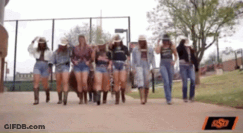 A Group Of Women Wearing Cowboy Hats And Shorts Walking Down A Sidewalk GIF