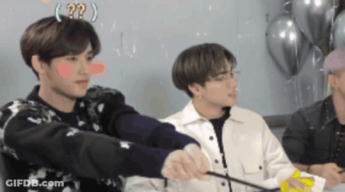 A Group Of Young Men Are Sitting Around A Table With Balloons In The Background And One Of Them Has A Question Mark On H GIF