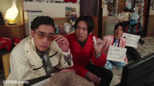 A Group Of Young Men Are Sitting In Front Of A Television And One Of Them Is Holding A Sign That Says ' A ' GIF