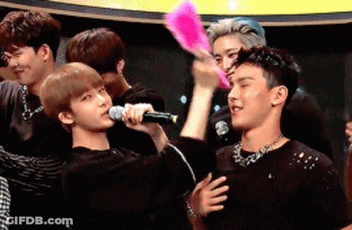 A Group Of Young Men Are Standing Around A Man Holding A Microphone And A Fan GIF
