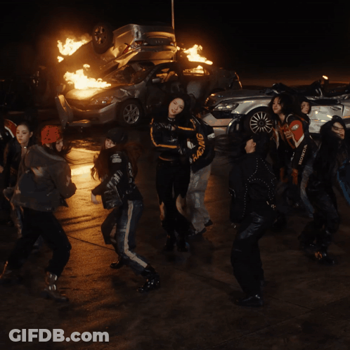 A Group Of Young Women Are Dancing In Front Of A Pile Of Burning Cars GIF