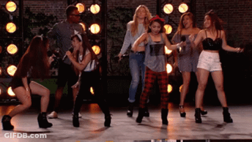 A Group Of Young Women Are Dancing On A Stage With A Man Holding A Microphone GIF