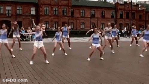 A Group Of Young Women Are Dancing On A Wooden Floor In Front Of A Brick Building . GIF