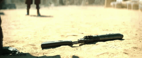 A Gun Is Laying In The Sand With A Person Walking In The Background GIF