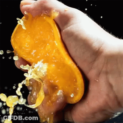 A Hand Is Squeezing A Slice Of Orange With Juice Splashing Out Of It GIF